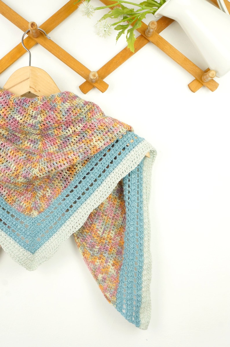 May include: A colorful, handmade crochet shawl in pastel shades of pink, yellow, and blue. The shawl is triangular, with a light blue border and a decorative openwork design. It hangs on a wooden hanger against a white background.