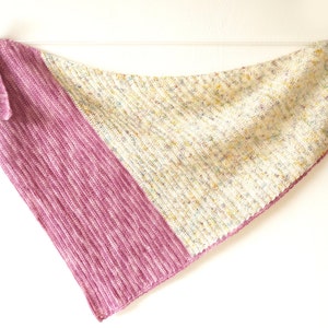 K&ouml;nnte beinhalten: Ein dreieckiger gestrickter Schal mit einem rosa-wei&szlig; gesprenkelten Muster. Der Schal h&auml;ngt an zwei W&auml;scheklammern vor einem wei&szlig;en Hintergrund.