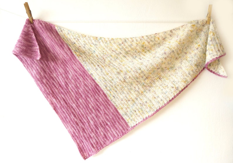 K&ouml;nnte beinhalten: Ein dreieckiger gestrickter Schal mit einem rosa-wei&szlig; gesprenkelten Muster. Der Schal h&auml;ngt an zwei W&auml;scheklammern vor einem wei&szlig;en Hintergrund.