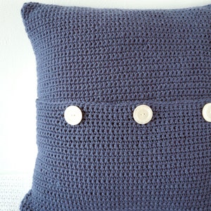 Puede incluir: Una almohada azul de crochet con tres botones blancos. La almohada est&aacute; hecha con un punto texturizado.