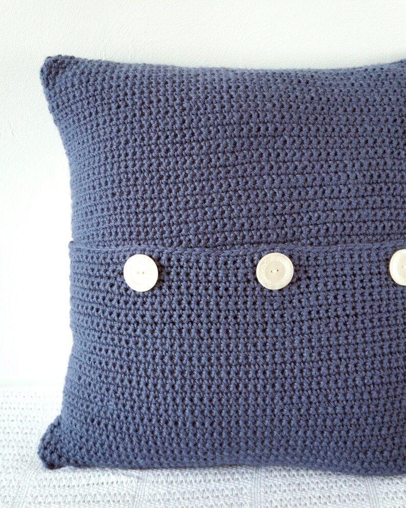 Puede incluir: Una almohada azul de crochet con tres botones blancos. La almohada est&aacute; hecha con un punto texturizado.