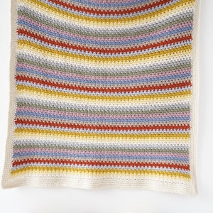 May include: A striped crocheted baby blanket in shades of yellow, orange, red, blue, green, and pink. The blanket is hanging on a white wall with a row of yarn balls in the same colors in front of it.