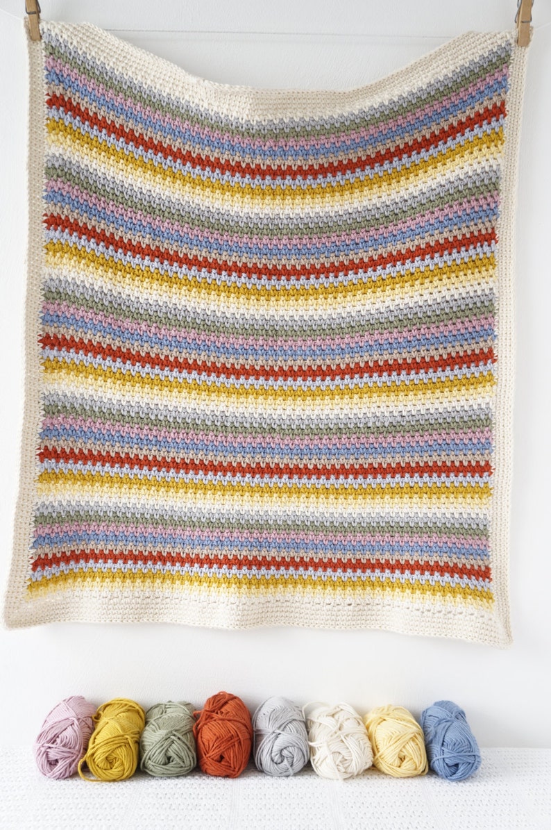 May include: A striped crocheted baby blanket in shades of yellow, orange, red, blue, green, and pink. The blanket is hanging on a white wall with a row of yarn balls in the same colors in front of it.