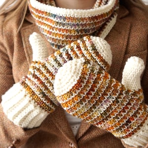 May include: A pair of crocheted fingerless gloves with a white cuff and a colorful striped pattern in shades of brown, orange, and cream. The gloves are worn by a person wearing a brown tweed jacket.