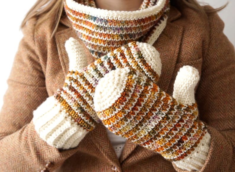 May include: A pair of crocheted fingerless gloves with a white cuff and a colorful striped pattern in shades of brown, orange, and cream. The gloves are worn by a person wearing a brown tweed jacket.