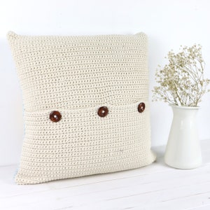 Puede incluir: Una almohada de crochet de color crema con tres botones marrones. La almohada est&aacute; sobre una superficie blanca.