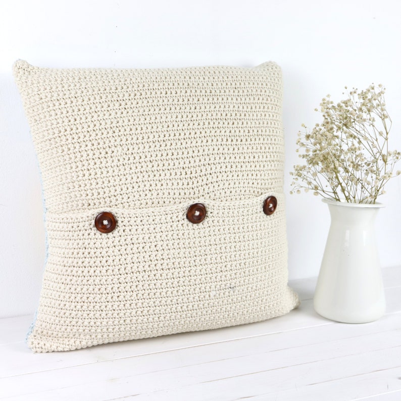 Puede incluir: Una almohada de crochet de color crema con tres botones marrones. La almohada est&aacute; sobre una superficie blanca.