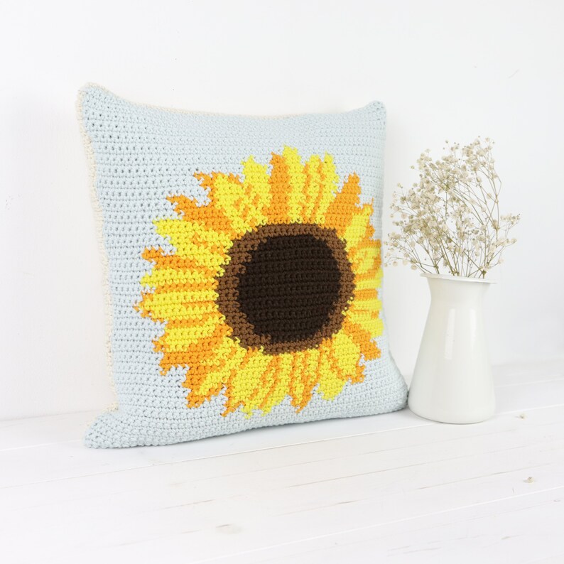 Puede incluir: Una almohada de crochet con un dise&ntilde;o de girasol. El girasol es amarillo y naranja con un centro marr&oacute;n. La almohada es azul claro y tiene un borde blanco.