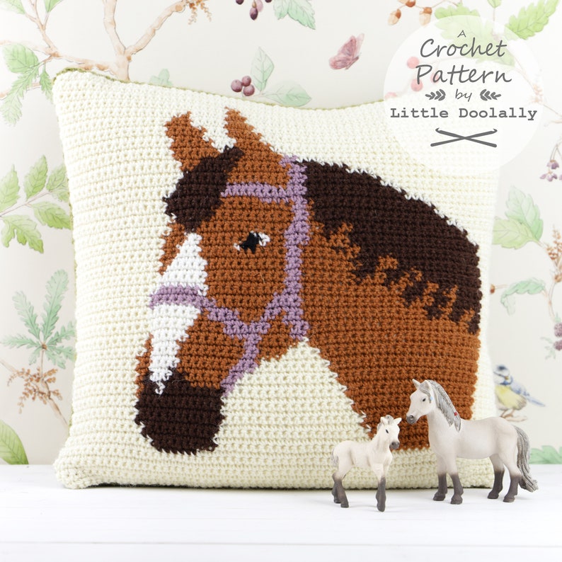 May include: A crochet cushion featuring a brown horse head design. The cushion is cream coloured with a brown, white and purple horse head in the centre. The horse is wearing a purple bridle. Two small toy horses are in the foreground.