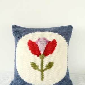 Puede incluir: Una almohada de punto de crochet azul y blanco con un dise&ntilde;o de flor roja, rosa y verde en el centro.