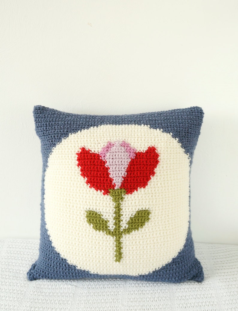 Puede incluir: Una almohada de punto de crochet azul y blanco con un dise&ntilde;o de flor roja, rosa y verde en el centro.