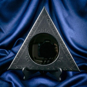 May include: A triangular black object with a circular center cutout. The surface features embossed text including the words TETRAGRAMMATON, ANAPHAXETON, PRIMEUMATON, and CHA MI. The object is supported by a small black stand at the base.