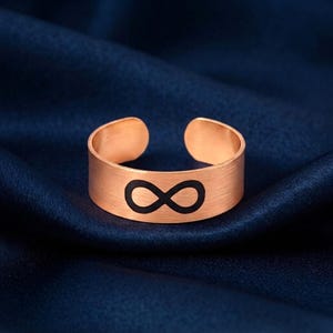 May include: A rose gold-toned open band ring featuring a brushed metal finish and a black infinity symbol engraved in the center. The ring has a wide, flat profile with a gap at the back, allowing for adjustable sizing to fit various finger widths.