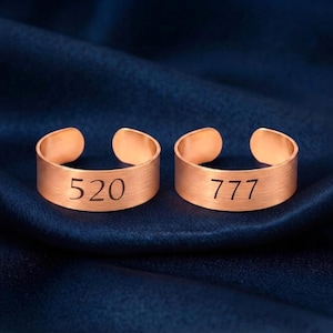 May include: Two rose gold-toned open rings with a brushed metal finish. The left ring is engraved with the number 520, and the right ring is engraved with the number 777. Both rings feature a simple band design with a gap at the back.