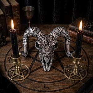 May include: A silver-toned ram skull sculpture featuring intricate filigree and Celtic knotwork patterns, positioned between two black candles in brass pentagram-shaped candle holders on a circular wooden board with a carved pentagram design.