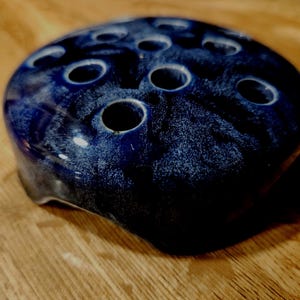 May include: A dark blue ceramic flower frog with multiple holes for arranging stems. The frog has a speckled, glossy finish and sits on a wooden surface. The holes are white-rimmed, and the overall shape is rounded.
