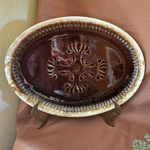May include: An oval ceramic platter featuring a deep brown glazed center with a symmetrical, embossed floral-like pattern. The rim is textured with a repeating vertical ridge design, finished in a contrasting cream and dark brown drip glaze.