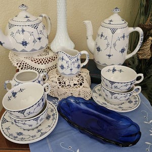 May include: A collection of white porcelain teacups, saucers, teapots, a creamer, and a sugar bowl, all with a blue floral design. A cobalt blue glass serving dish is also present. The tea set is arranged on a table with lace doilies.