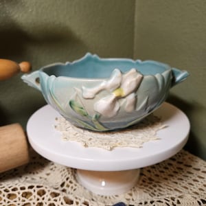 May include: A teal ceramic bowl featuring a raised, sculpted white iris flower with a yellow center on the exterior. The bowl has a scalloped rim and two small handles on opposite sides, with a smooth, matte finish and subtle green leaf accents.