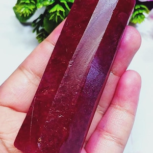Puede incluir: Un espécimen de cristal hexagonal alargado de color carmesí profundo con una superficie pulida y facetada. La piedra muestra un tono rojo rico y saturado con variaciones sutiles en la textura y claridad a lo largo de su estructura prismática.