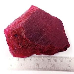 May include: A rough, uncut ruby gemstone with a deep red hue. The stone has a textured surface and irregular shape. A metal ruler is placed next to the stone, showing a length of approximately 4 inches.