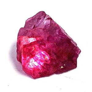 Könnte beinhalten: Ein roher, ungeschliffener Edelstein in tiefem Magenta und leuchtendem Pink. Der Stein weist eine gezackte, kristalline Struktur mit lichtdurchlässigen Bereichen auf, die das Licht einfangen und scharfe Kanten sowie eine raue Oberfläche zeigen.
