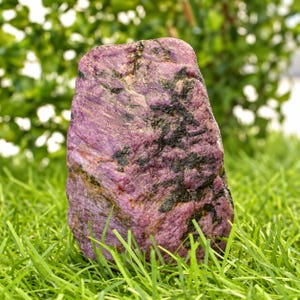 Peut inclure: Un spécimen de cristal de rubis brut et vertical présentant une teinte magenta et violette profonde avec des inclusions minérales noires proéminentes et une surface rugueuse et texturée. La pierre montre des formations cristallines naturelles.