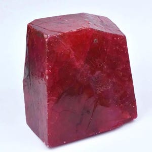 Può includere: Un cristallo di rubino grezzo rosso intenso con una struttura a blocchi sfaccettata. La pietra mostra una qualità traslucida con fratture interne visibili, texture superficiali naturali e un aspetto lucido sulle sue facce principali.