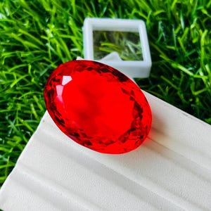 May include: A vibrant, oval-shaped red gemstone with a faceted surface that reflects light. The gemstone is positioned on a white, ridged display stand, with a small, square white box visible in the background behind the stone.