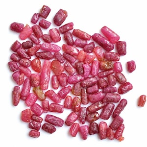 Può includere: Una collezione di piccole pietre preziose di rubino grezze e cilindriche in varie tonalità di rosa intenso e rosso. Ogni pietra presenta una consistenza naturale non lucidata con sfaccettature cristalline visibili e superfici irregolari.