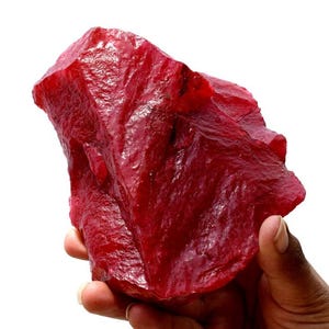May include: A large, raw, deep red gemstone with a jagged, crystalline surface texture. The stone features irregular facets and sharp edges, displaying a vibrant, saturated crimson color throughout its translucent and opaque sections.