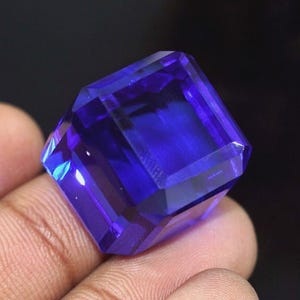May include: A faceted, deep blue gemstone with an emerald cut. The gem is held in a hand, and the facets reflect light, creating highlights. The stone's colour is a rich, saturated blue, and the cut is precise.
