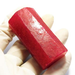 May include: A faceted, rectangular ruby gemstone with a deep red color. The gemstone is held in a gloved hand against a white background. The ruby's surface has a slightly rough texture, and its shape is a long, hexagonal prism.