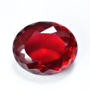 May include: A faceted, oval-shaped, deep red gemstone. The gem has a brilliant, polished surface with multiple facets that reflect light. The color is a rich, saturated red, with a slight variation in tone across the surface.
