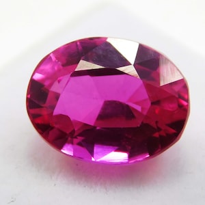Puede incluir: Una gema facetada de forma ovalada con un tono magenta intenso. La superficie de la gema refleja la luz, mostrando sus múltiples facetas y estructura interna. La piedra parece ser un rubí, una gema preciosa que se usa a menudo en joyería.