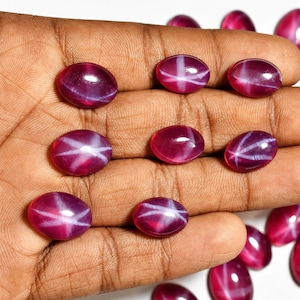 May include: A collection of oval-shaped, deep red gemstones with a star-like white pattern. The gems are smooth and polished, reflecting light. The stones are held in a hand, showcasing their color and design. These gemstones are likely used for jewelry.