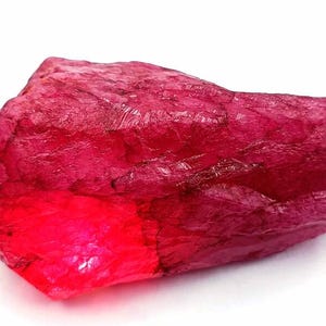 May include: A raw, unpolished ruby gemstone with a deep, vibrant magenta-red colour. The stone features a rough, crystalline texture with natural facets and internal light reflections that create varying shades of intense red across its surface.