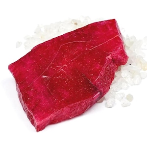 Pode incluir: Uma grande pedra preciosa de rubi bruta de cor vermelho-profundo, com uma forma irregular facetada e estrias superficiais visíveis, repousando sobre uma cama de pequenos fragmentos minerais cristalinos brancos translúcidos.