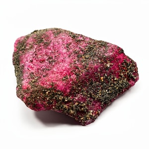 May include: A rough, uncut ruby gemstone with a vibrant pinkish-red hue. The surface is textured with dark, speckled inclusions, creating a contrast against the bright color. The gemstone is displayed against a white background.