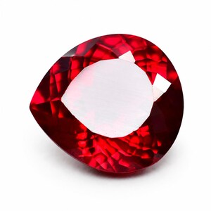May include: A faceted, pear-shaped ruby gemstone. The gem is a deep, saturated red with a bright, reflective surface. The cut reveals internal facets, enhancing its brilliance. The gem is set against a white background, highlighting its colour and shape.
