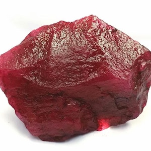 Puede incluir: Una piedra preciosa de rubí en bruto y sin pulir con un color rojo carmesí profundo y saturado. La piedra presenta una forma irregular y dentada con una superficie cristalina texturizada que muestra niveles variables de translucidez.