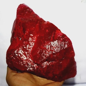 May include: A large, raw, deep red gemstone with a crystalline, irregular, and faceted surface texture. The stone displays varying shades of dark crimson and ruby red, with reflective highlights across its uneven, jagged, and natural rock-like form.
