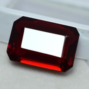 Peut inclure: Une pierre précieuse rectangulaire et facettée d'un rouge profond. La gemme présente un centre clair et rectangulaire et des bords biseautés, créant un contraste saisissant. La pierre est probablement un rubis ou un grenat.