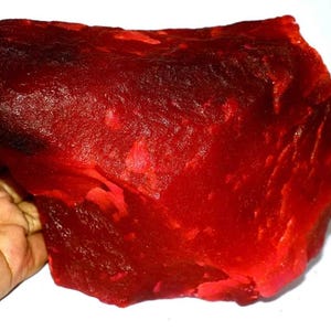 Könnte beinhalten: Ein großer, roher, unpolierter Brocken aus tiefrotem Mineral mit einer durchscheinenden, kristallinen Textur. Der Stein weist unregelmäßige, gezackte Kanten und eine raue, unebene Oberfläche mit verschiedenen Schattierungen von leuchtendem Karmesinrot auf.