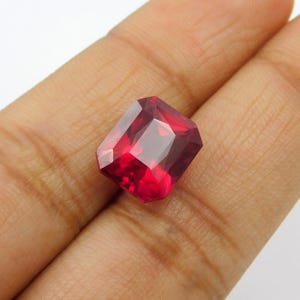 May include: A rectangular-cut ruby gemstone, displaying a rich, deep red hue. The gem features multiple facets, creating a sparkling effect. The ruby is held in a hand, highlighting its clarity and the quality of the cut.