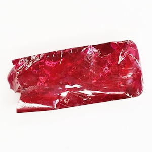 May include: A raw, uncut ruby gemstone with a deep red hue. The stone has a rough, irregular shape with visible fractures and a glossy surface. The ruby is set against a white background.