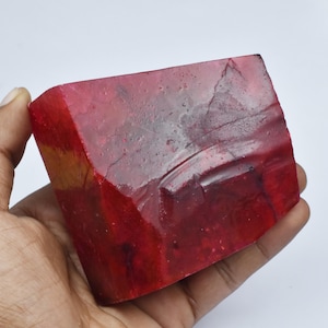 May include: A large, raw ruby gemstone with a deep red color. The ruby has a rectangular shape with visible natural textures and imperfections. The gemstone is held in a hand, showcasing its size and color.