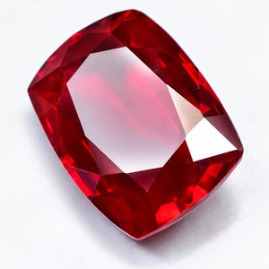 May include: A cushion-cut ruby gemstone with a deep, vibrant red colour and multiple triangular and trapezoidal facets. The stone exhibits high clarity and internal light reflection, creating a brilliant, polished appearance with sharp edges.
