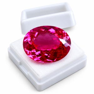 Puede incluir: Una gema de corte ovalado con un tono rosa magenta intenso y superficie facetada, que descansa dentro de una pequeña caja de presentación cuadrada de plástico blanco con la tapa abierta. La gema muestra gran claridad y refleja la luz.