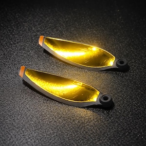 May include: Two gold-colored drone propellers with black and orange accents. The propellers have a curved, blade-like shape and are set against a dark, textured background. The propellers have a small, circular hole at the base.